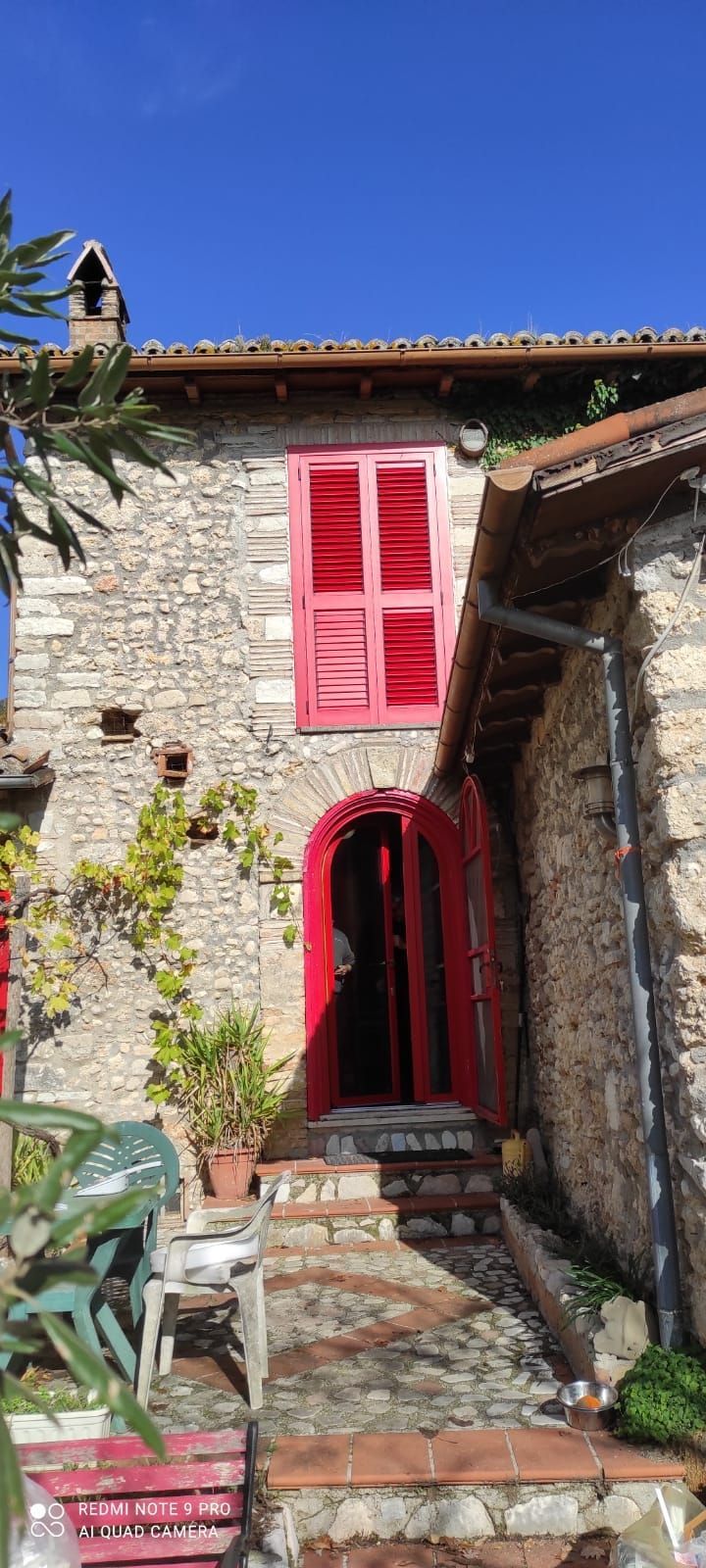 Una rustica casa in pietra con un portale ad arco rosso acceso, persiane rosse alla finestra superiore e un vialetto d'ingresso lastricato in pietra.