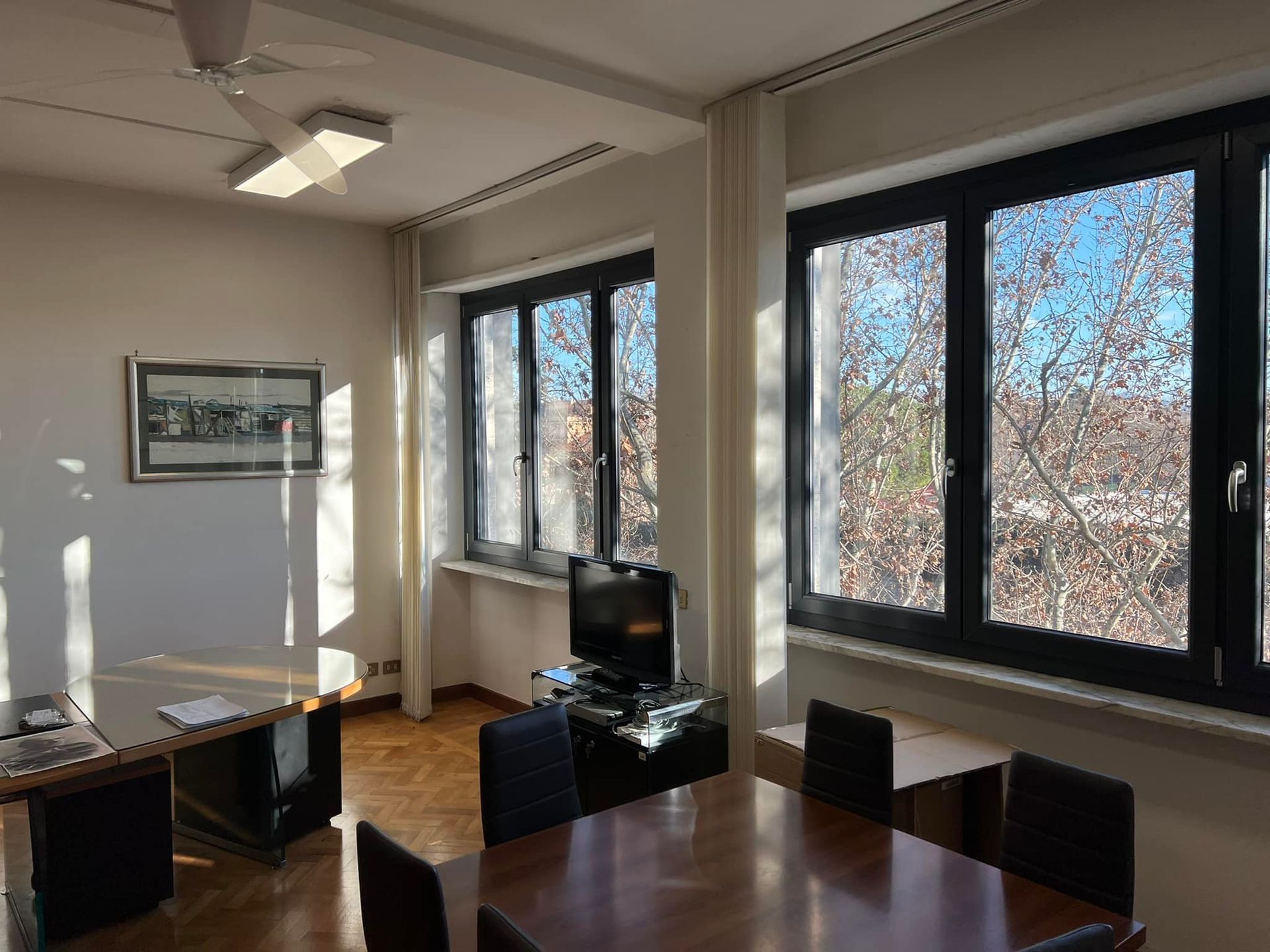 Un luminoso ufficio con un tavolo di legno, diverse sedie, una scrivania, un televisore e grandi finestre che si affacciano sugli alberi.