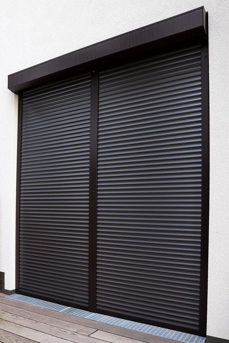 Persiane di sicurezza avvolgibili esterne di colore marrone scuro, installate sopra una porta su una parete bianca con finitura ruvida.