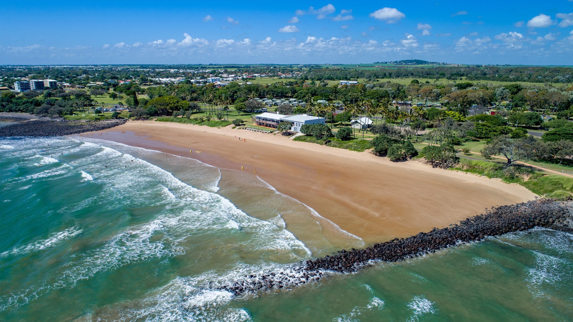 Aerial view of a sandy beach with turquoise water, a rocky breakwater, and buildings along the shore.