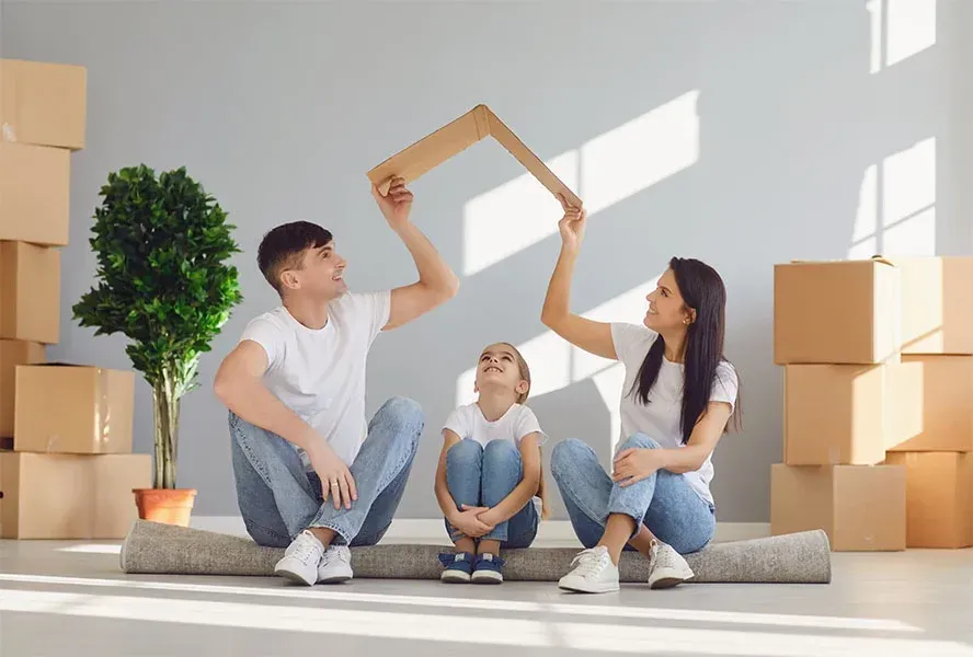 Family sitting, holding cardboard house shape, surrounded by moving boxes.