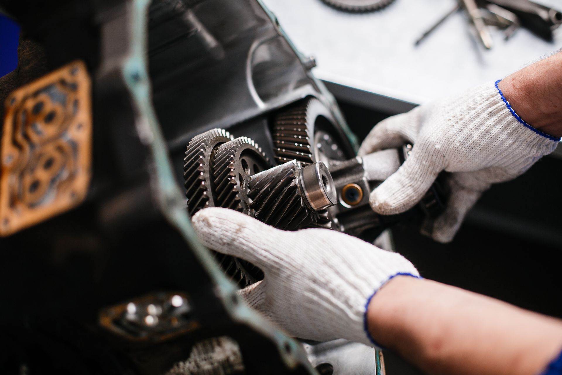 A mechanic wearing white work gloves holds and repairs a set of metal gears inside a car transmission.