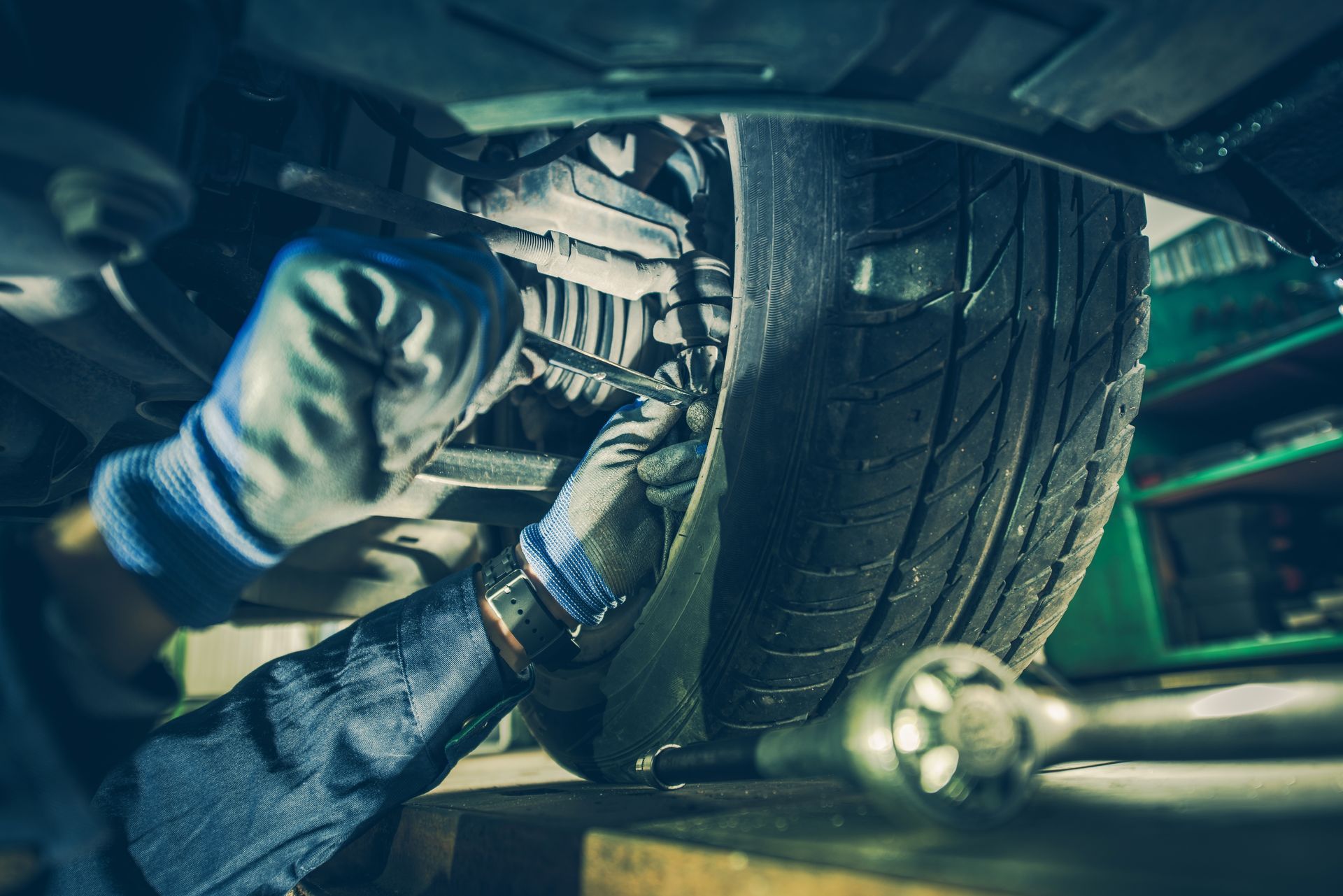 A mechanic in gloves uses tools to repair the suspension and tire assembly of a vehicle in a workshop.
