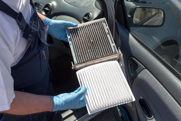 A mechanic in blue gloves holds a dirty car cabin air filter next to a new, clean replacement filter inside a car.