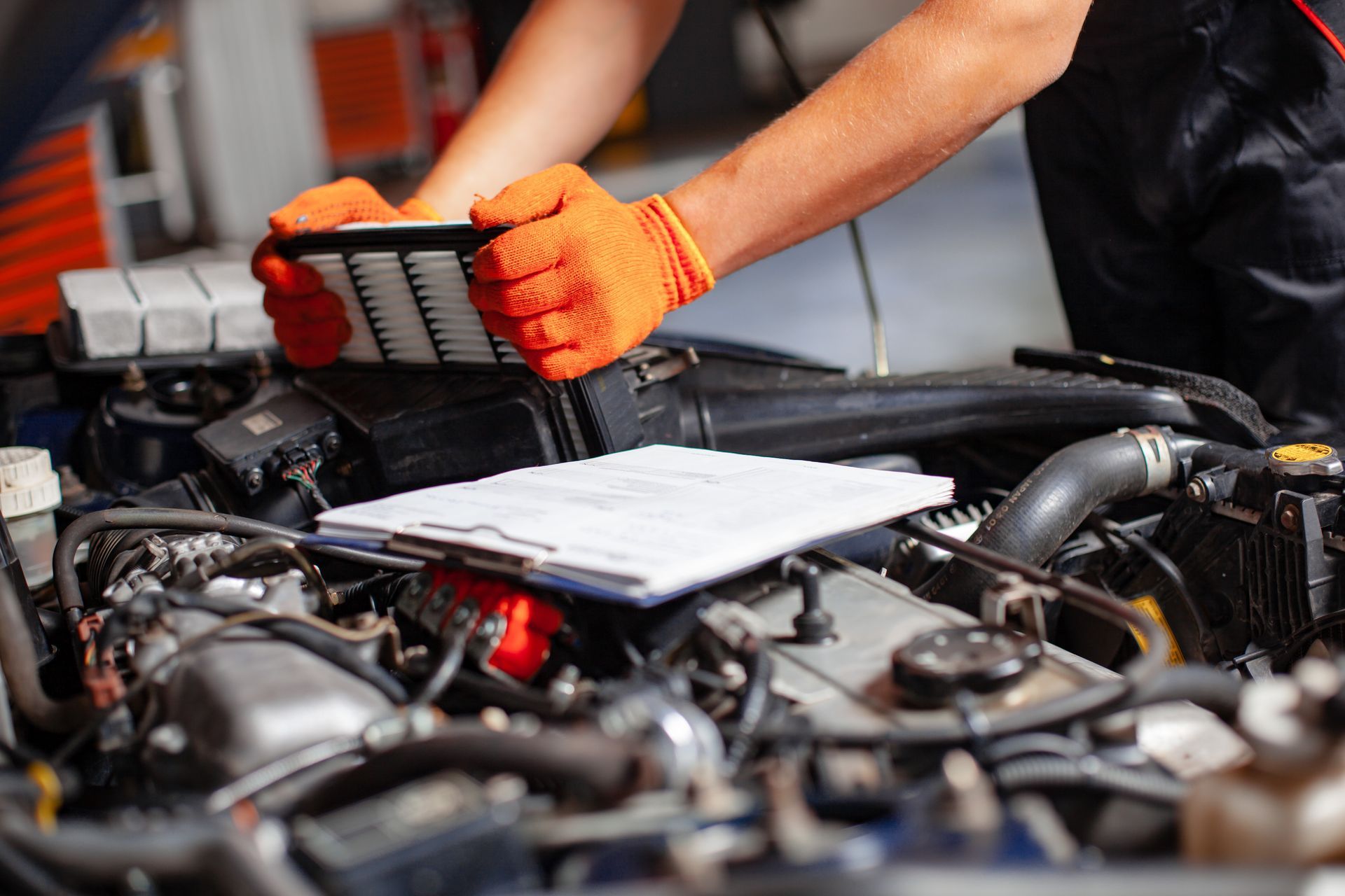A mechanic wearing orange gloves holds a rectangular air filter over an open car engine with a clipboard on top.