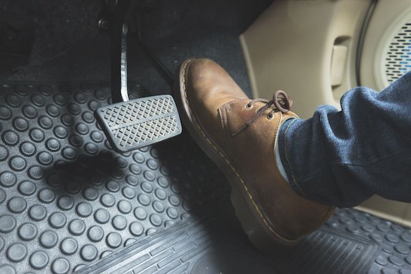 A person wearing a brown leather shoe rests their foot near a car's brake pedal above a circular textured floor mat.