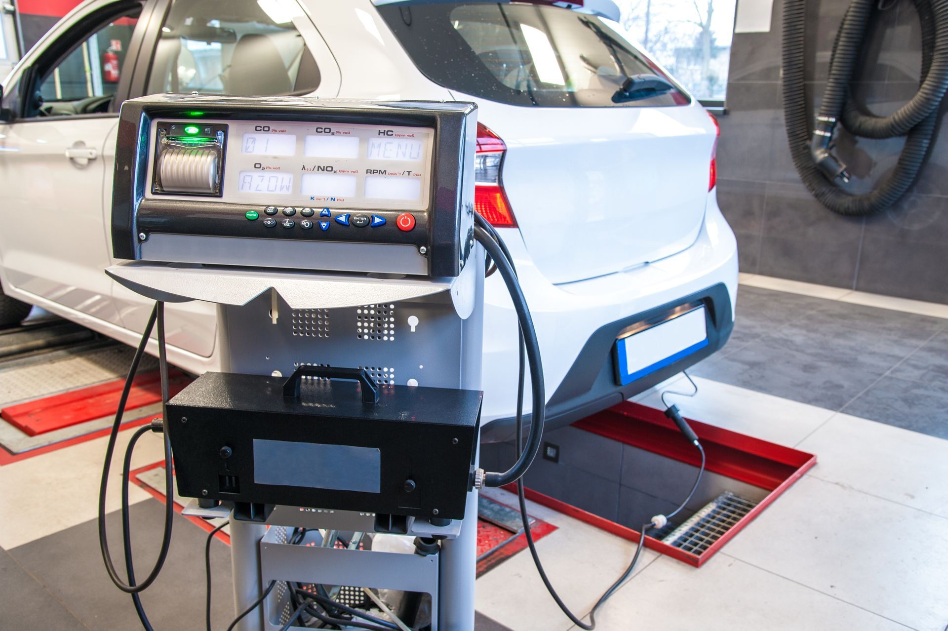 A white car undergoes an emissions test in a service garage, with testing equipment positioned in the foreground.