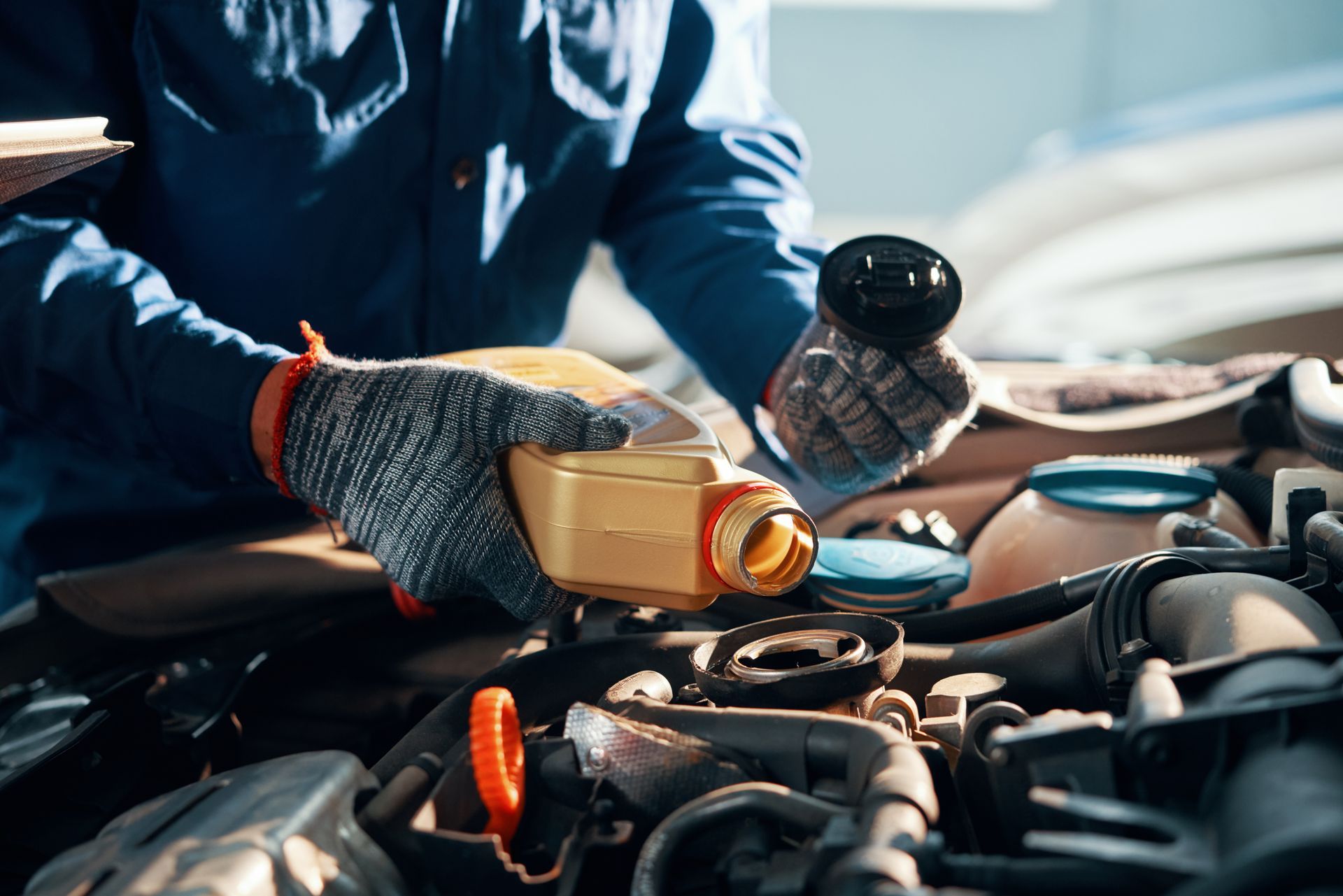 A mechanic in blue work clothes and gloves pours oil from a gold bottle into a car engine.