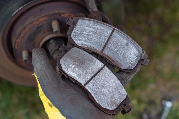 A person wearing a gloved hand holds two used, worn automotive brake pads in front of a rusty vehicle wheel hub.