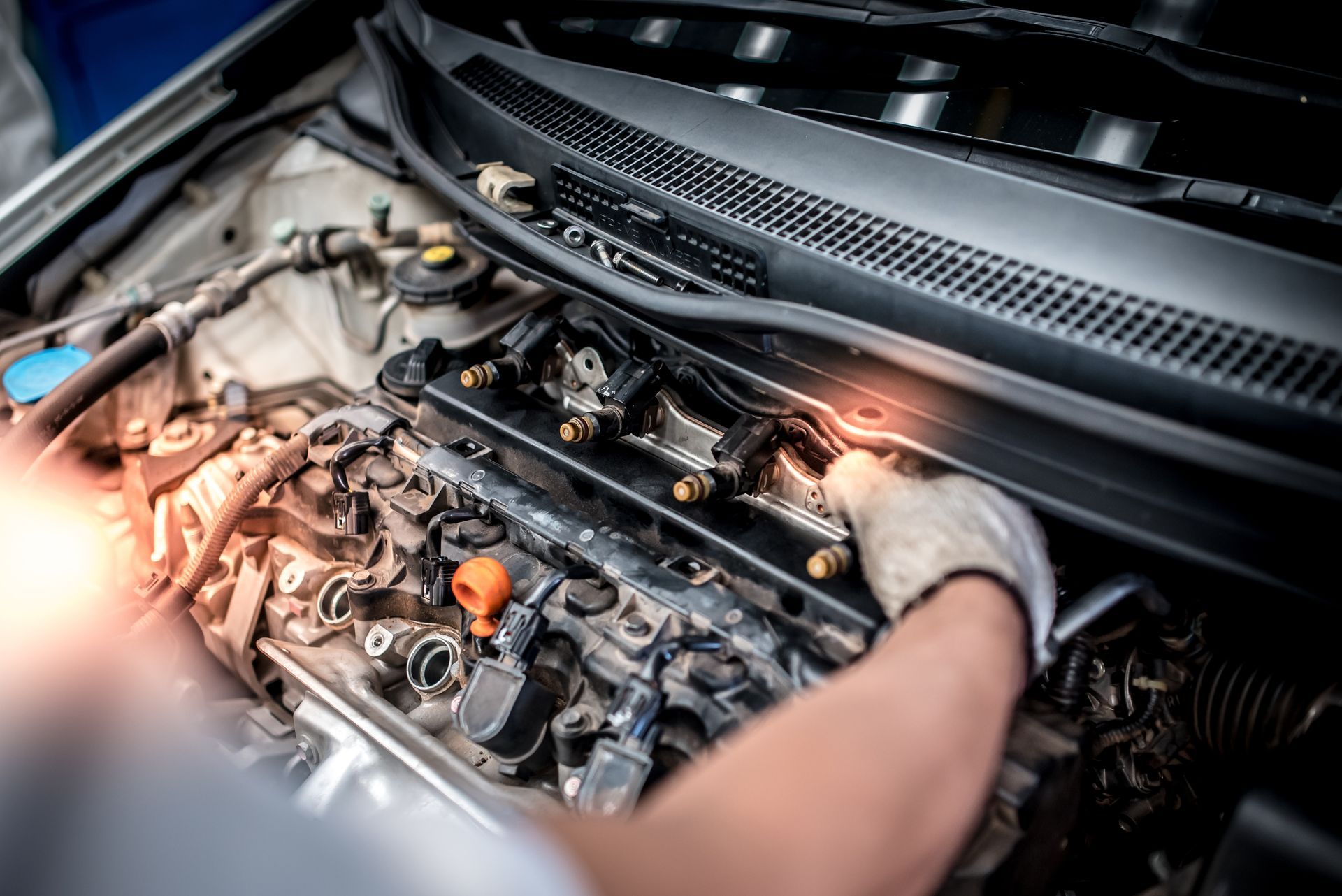 A mechanic’s gloved hand works on the fuel rail and engine components inside an open car hood.