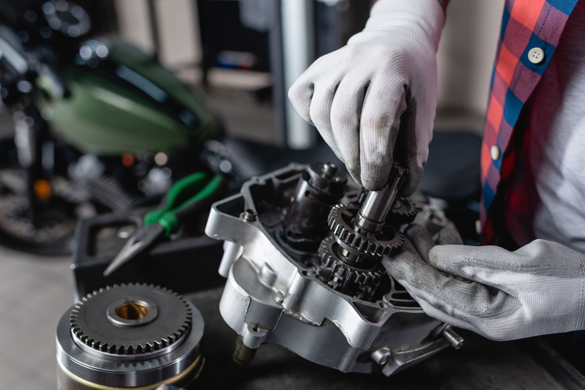 A mechanic in a plaid shirt wearing work gloves repairs a motorcycle engine component in a garage.