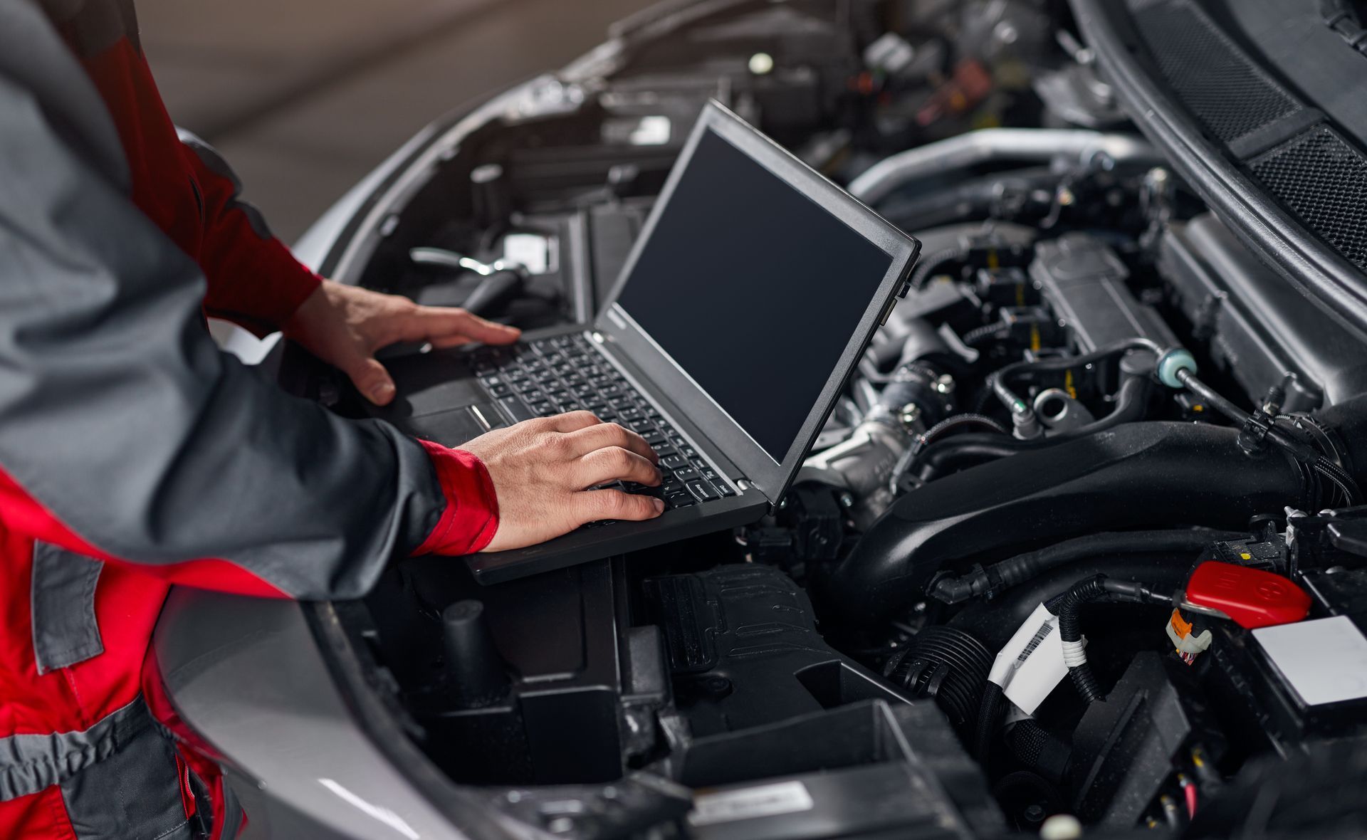A mechanic in a red and grey uniform uses a laptop to diagnose a car engine.