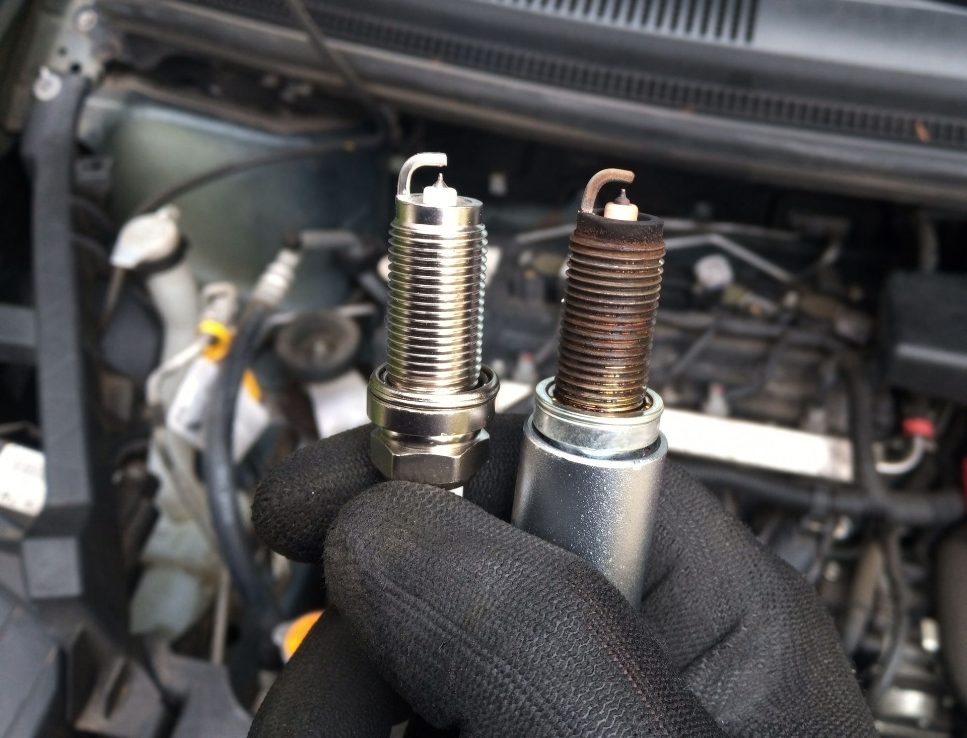 A gloved hand holds a new, clean spark plug next to a used, dirty one in front of an open car engine.