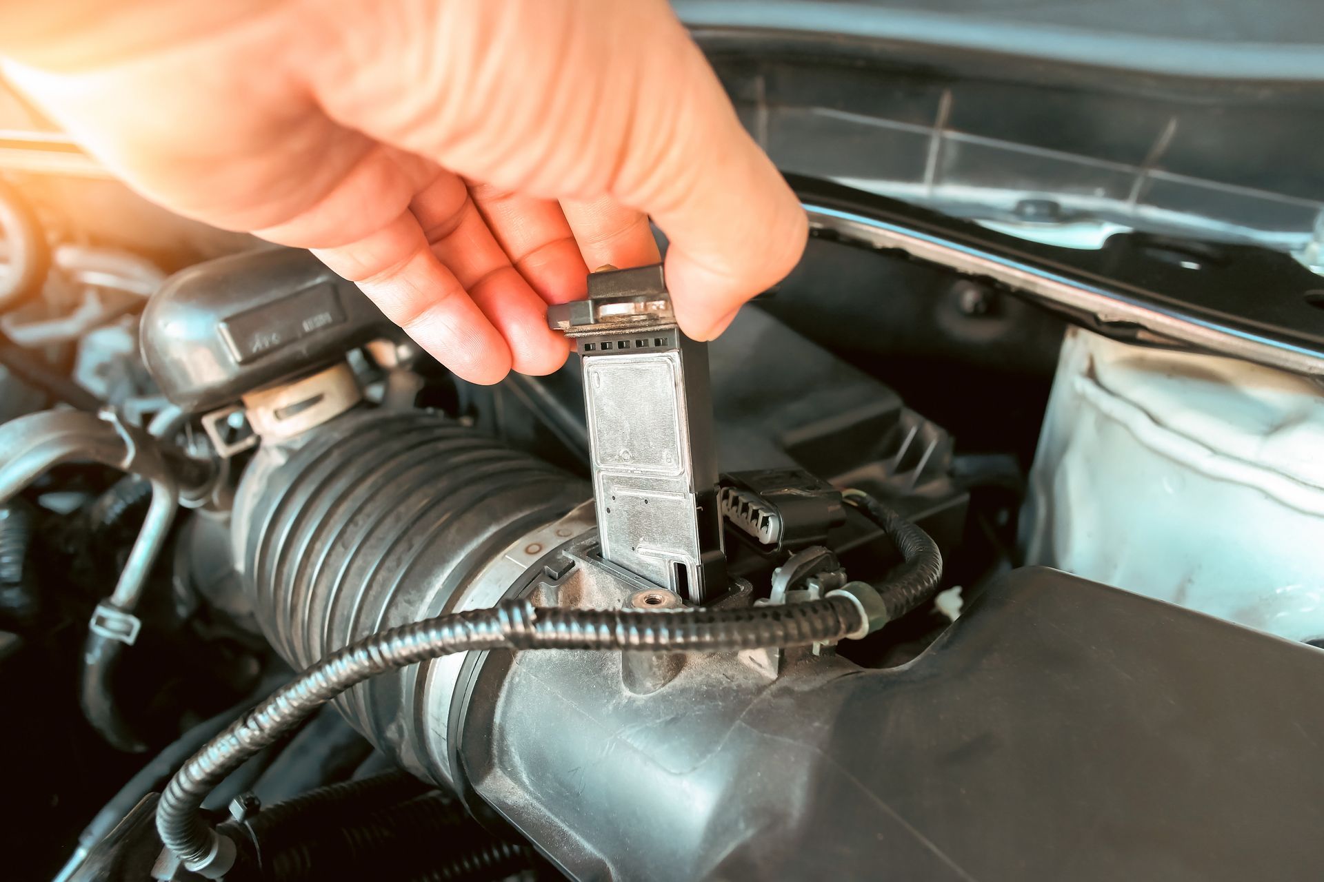 A hand removes a mass airflow sensor from a vehicle's air intake housing under the hood.