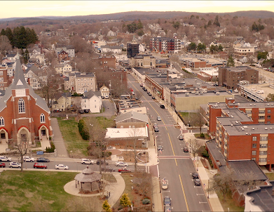 An aerial view of a town featuring a prominent red brick church, a gazebo in a park, and a main street with buildings.