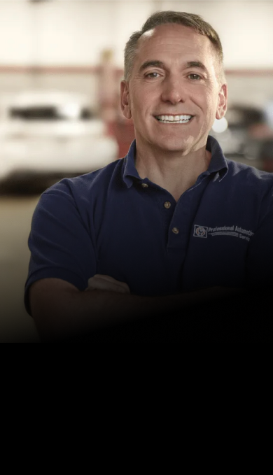 A smiling individual in a dark blue polo shirt stands outside a building with automotive signage in the background.