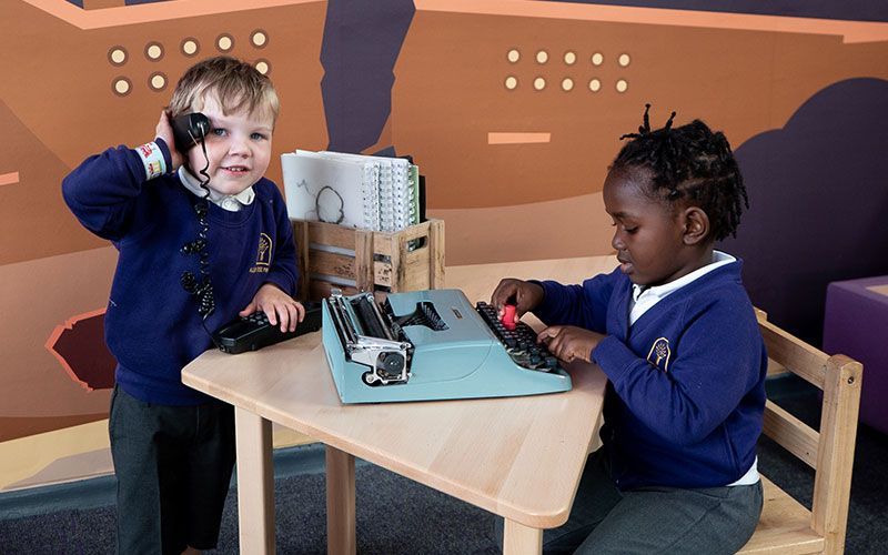 A boy and a girl are sitting at a table playing with a toy typewriter.