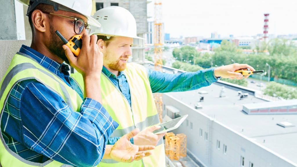 Two construction workers on a balcony, one using a walkie-talkie, the other pointing and holding a tablet.