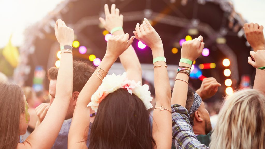 People with arms raised at an outdoor music festival with stage lights in the background.