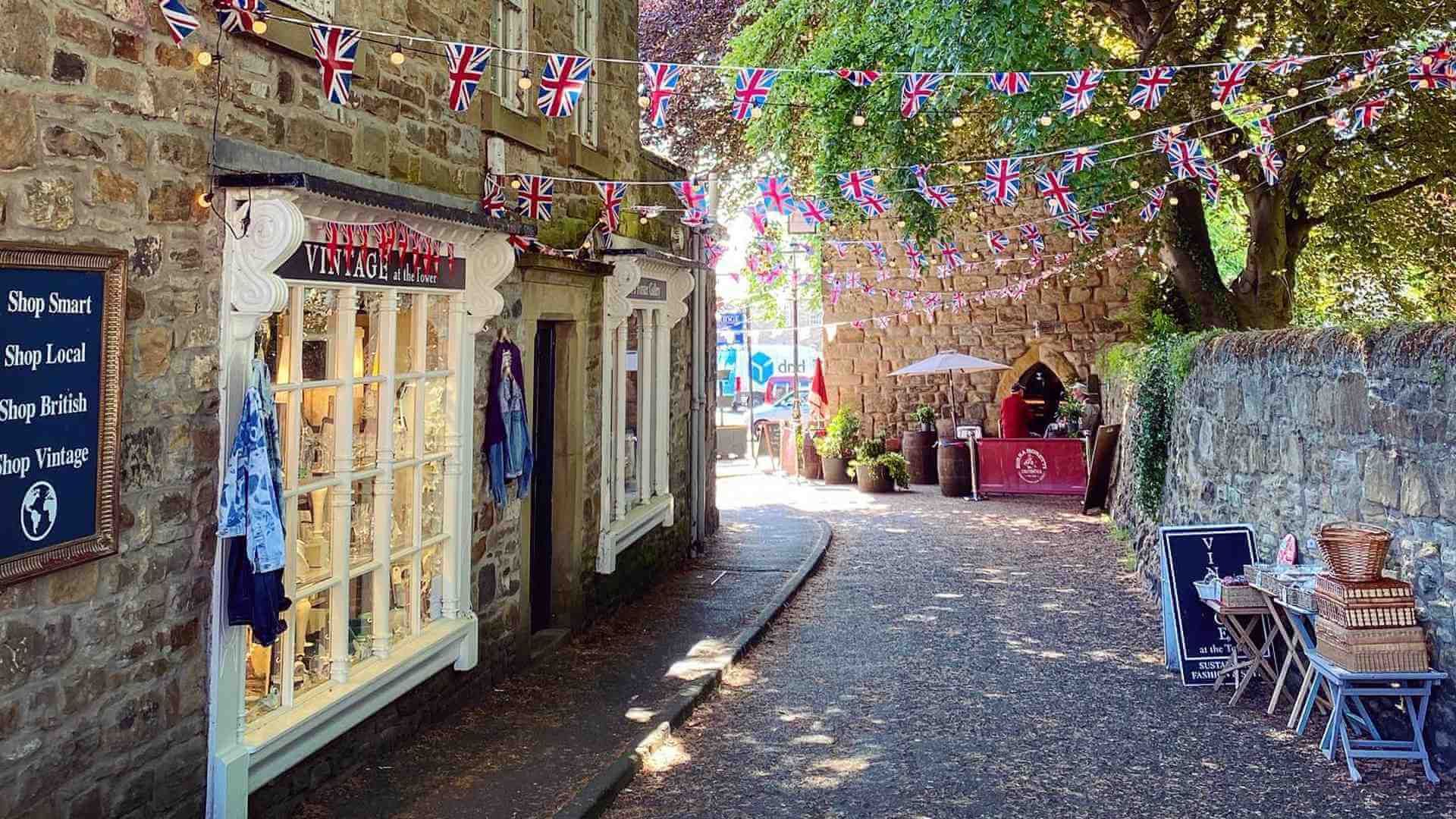 Corbirdge Quaint shop-lined lane with hanging flowers, Union Jack bunting, and a sunny cobblestone path