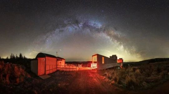 Night sky over lit buildings with a bright Milky Way arch above a rural landscape Kielder obs