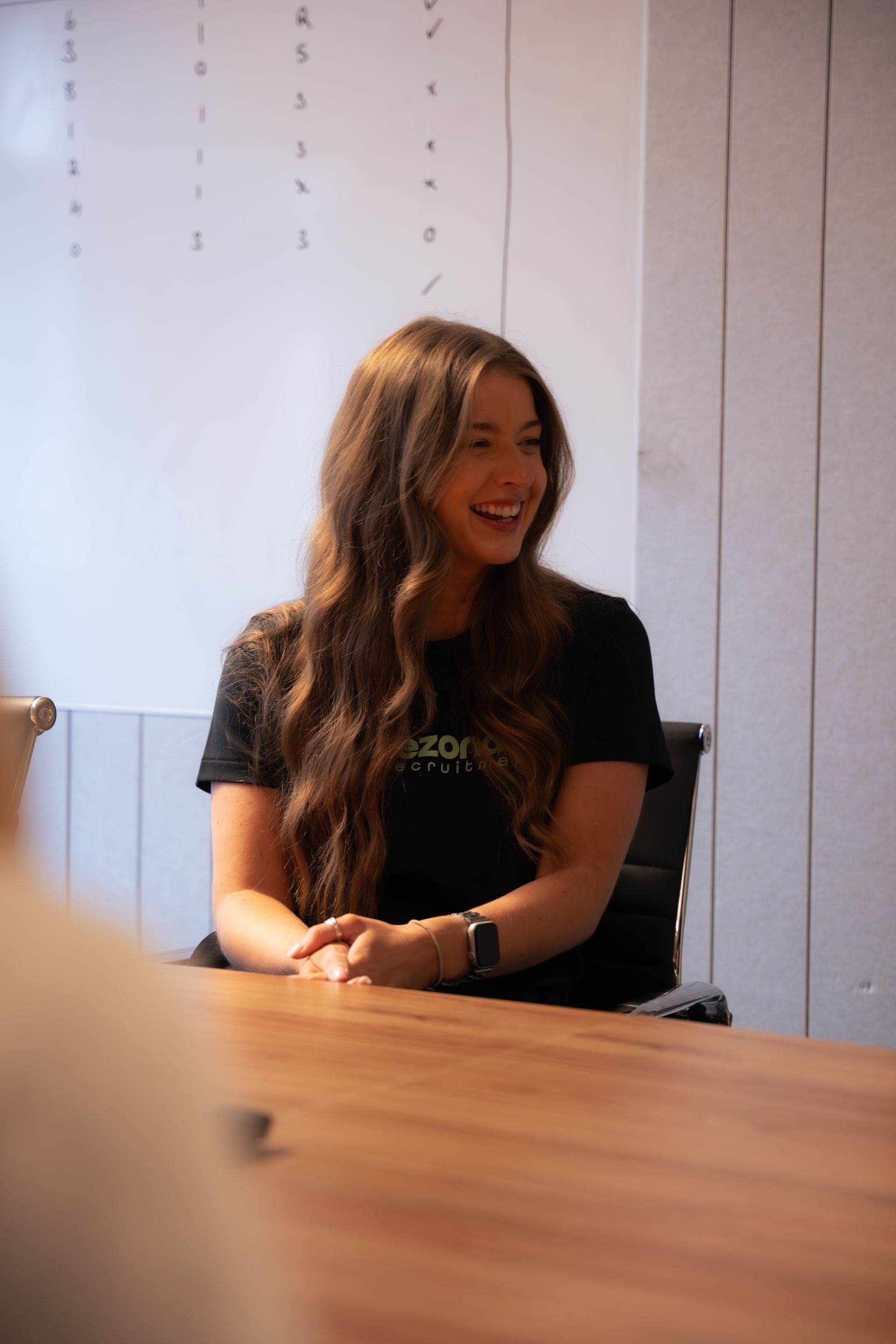 Woman wearing glasses and white blazer smiles while sitting at a laptop.