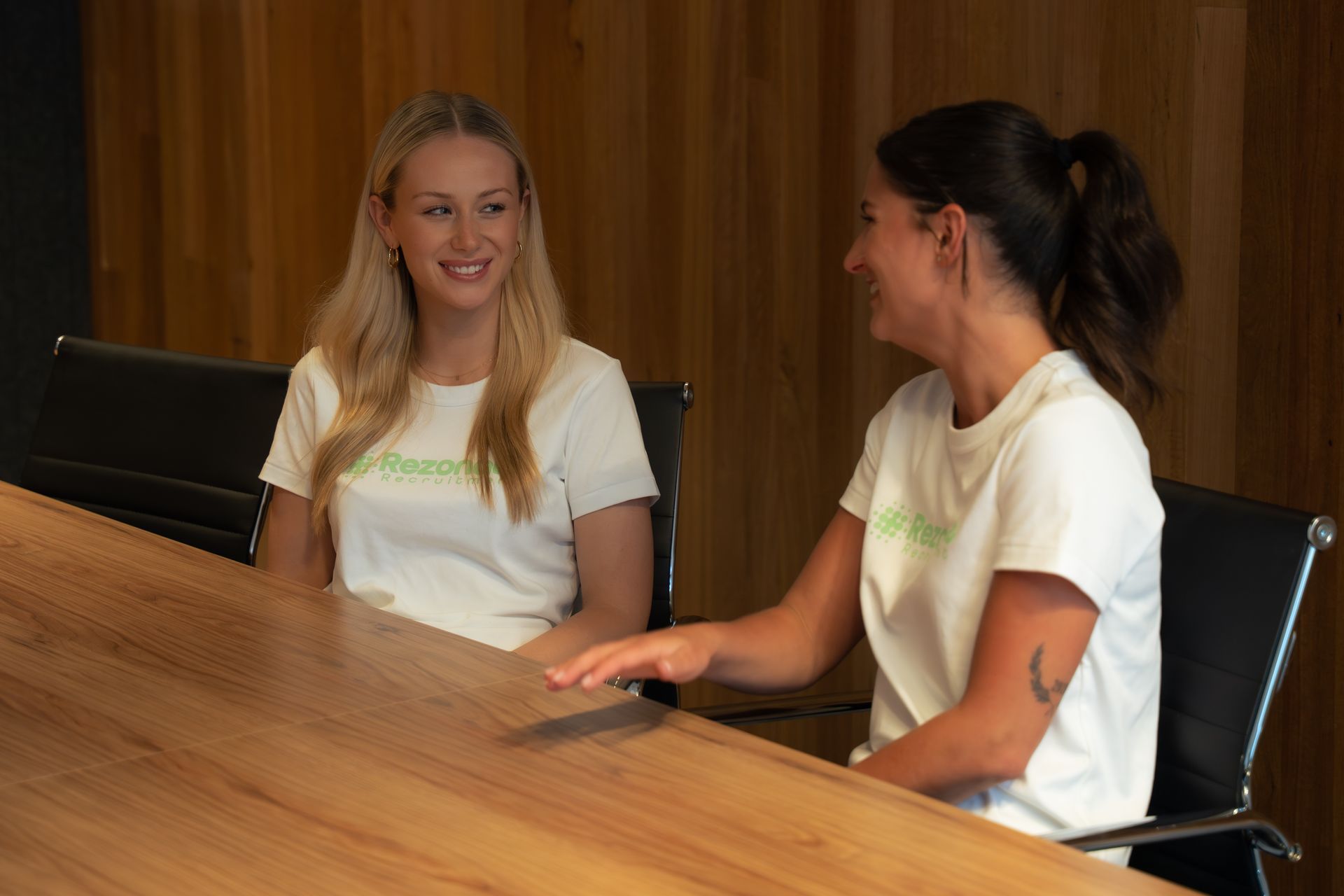 Woman in white blazer laughs, sitting at desk with laptop and glass. Bright room with window.