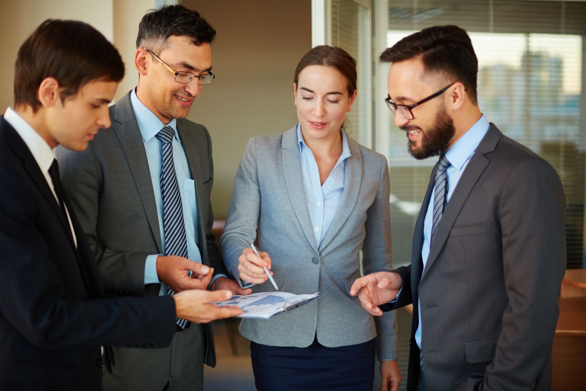 Four businesspeople in suits examining a document together; indoor office setting.