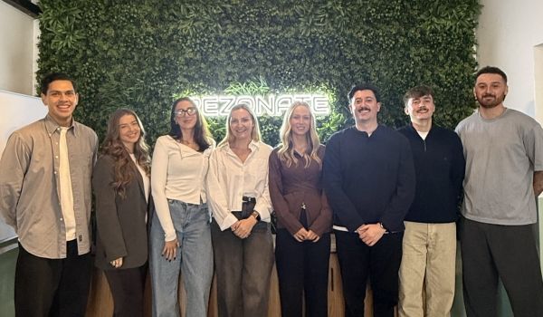 Eight people standing indoors in front of a green plant wall, posing for a group photo.