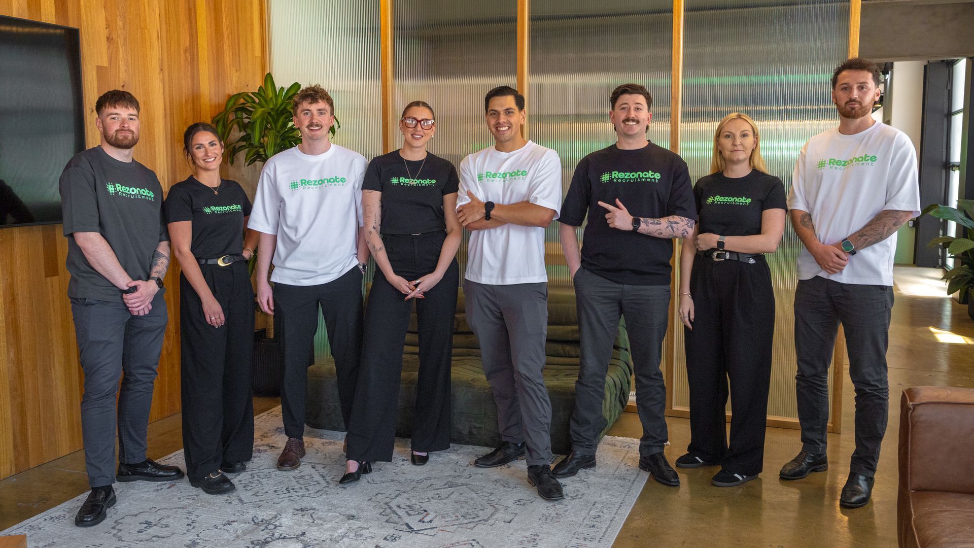 Woman in white blazer laughs, sitting at desk with laptop and glass. Bright room with window.