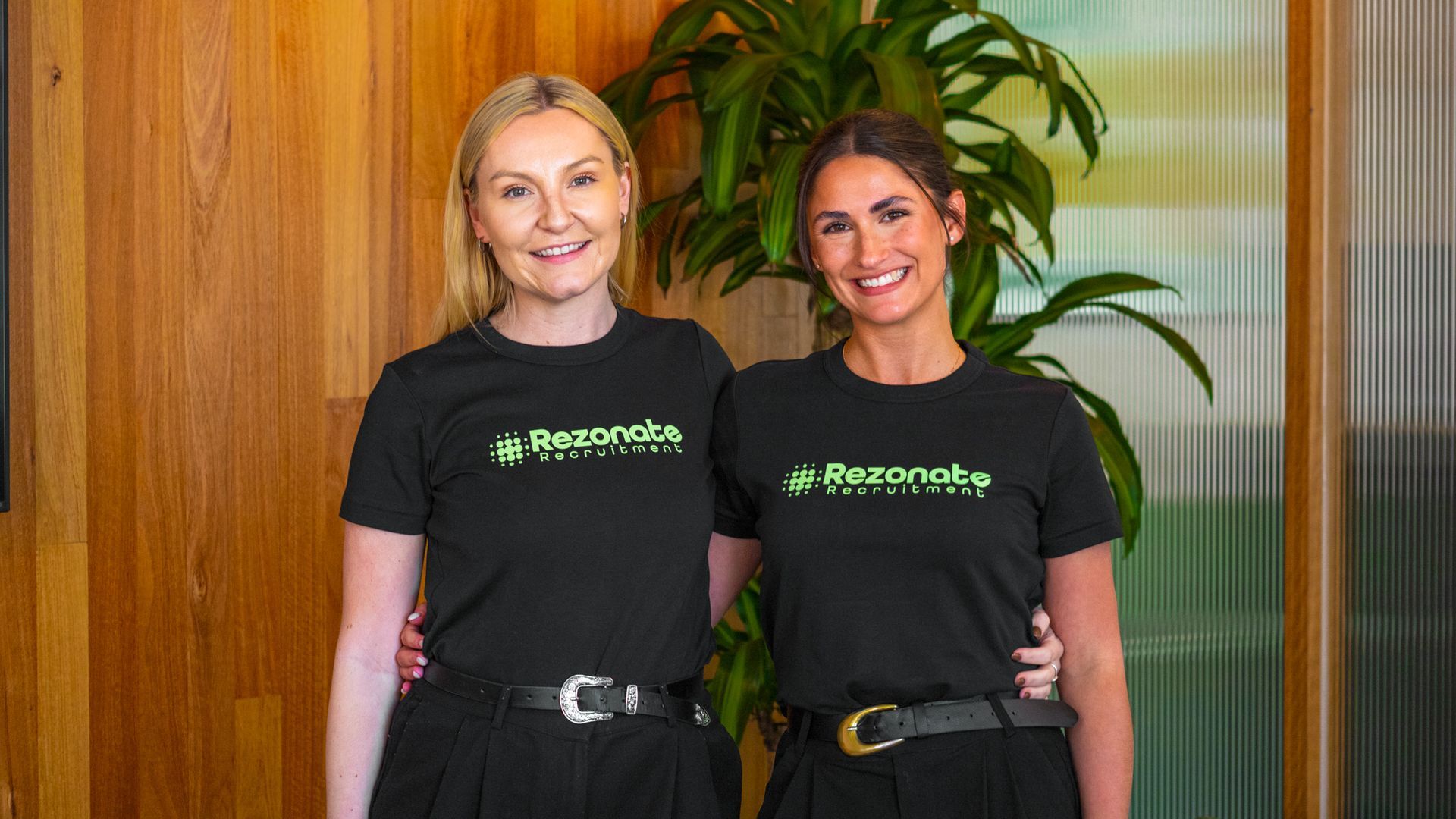 Woman wearing glasses and white blazer smiles while sitting at a laptop.