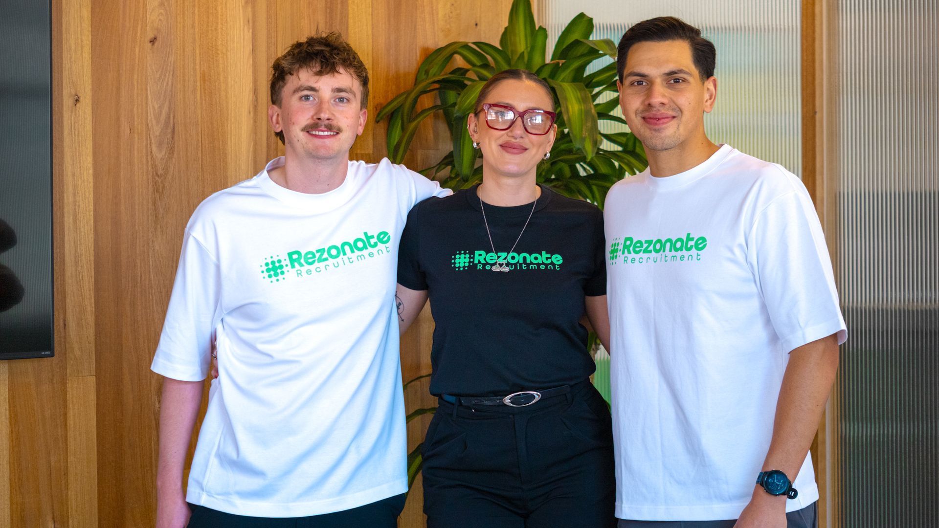 Three people stand together, smiling. They wear shirts with a green logo. Indoor setting, with a plant in the background.