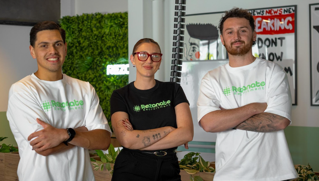 Group of people around a wooden table, two shaking hands, laptops and tablets visible.