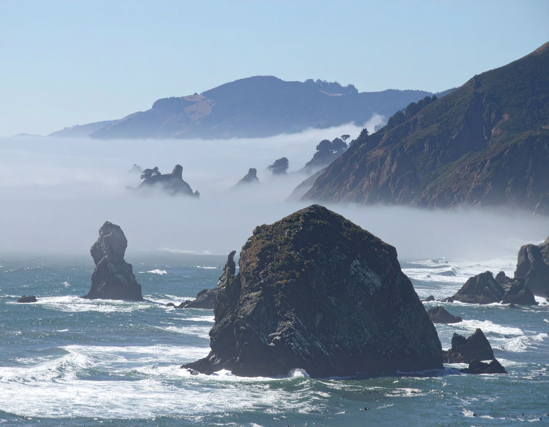 Rocky coastal shoreline with mist, ocean waves, and distant cliffs under a pale blue sky