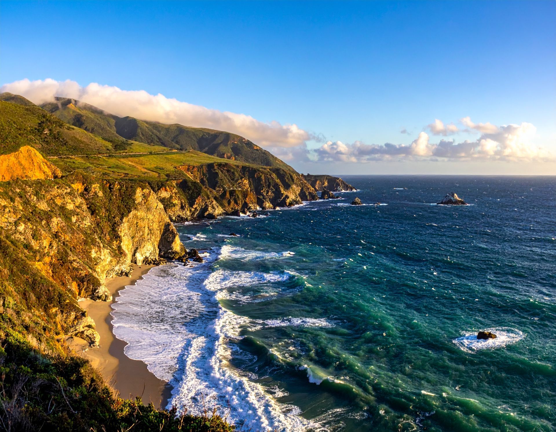 Rocky coastal cliffs beside a turquoise ocean with waves, sunlight, and a sandy cove
