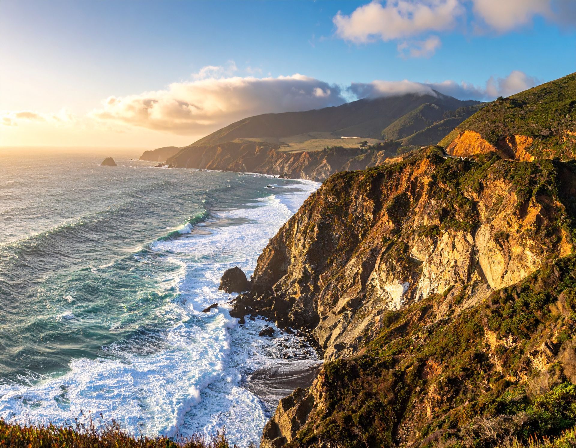 Sunlit coastal cliffs above a foamy ocean at sunset, with mountains in the distance