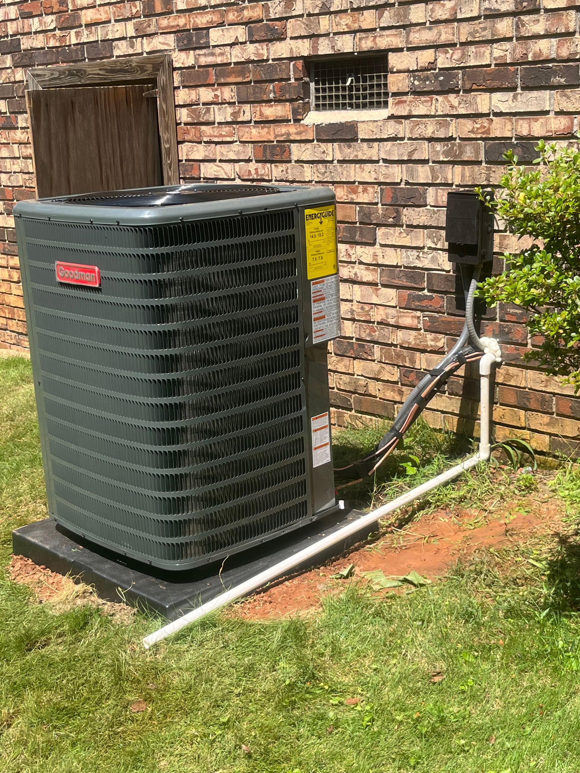 Central air conditioner unit outside, beside brick building, with white pipe leading from it.