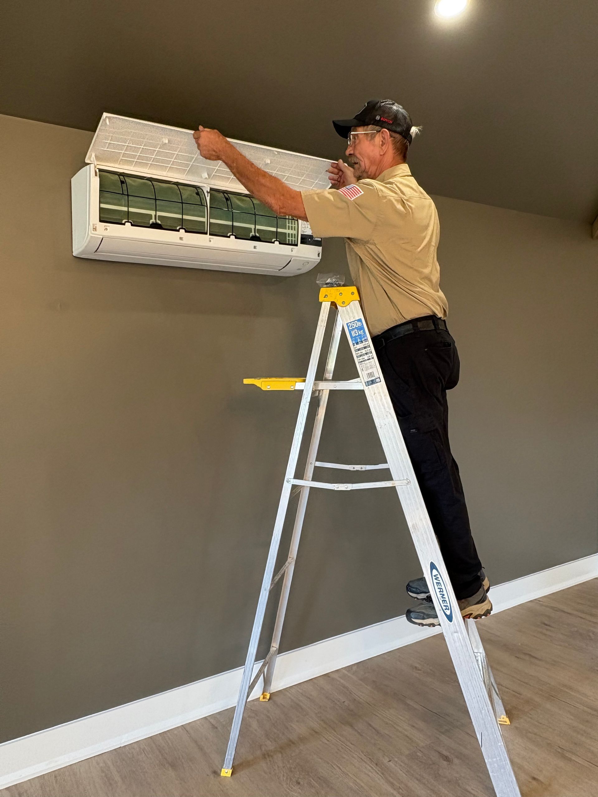 Man in uniform on a ladder cleaning a wall-mounted air conditioning unit indoors.