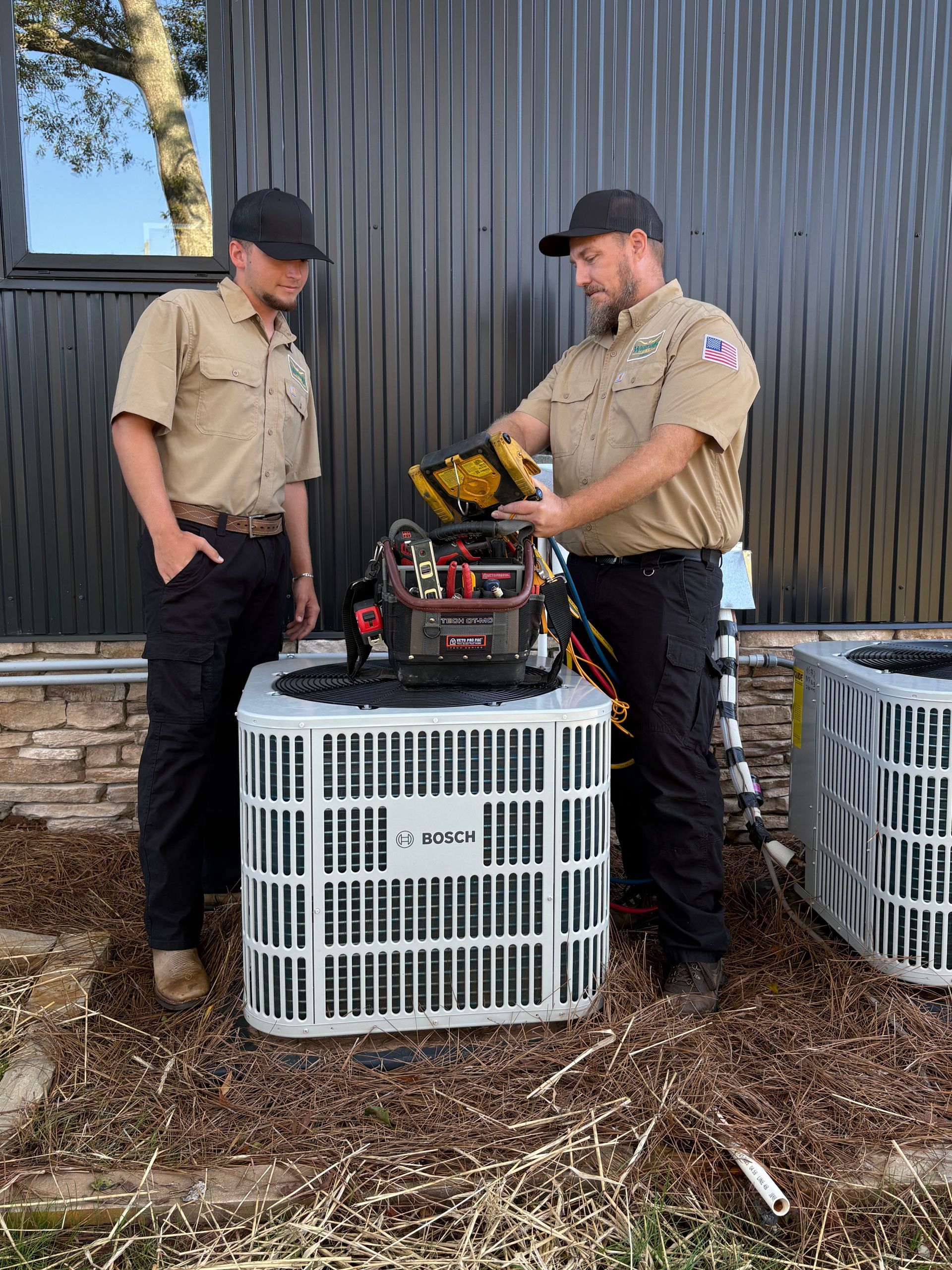 Two HVAC technicians inspecting an air conditioning unit outside a building; one holding a tool kit.