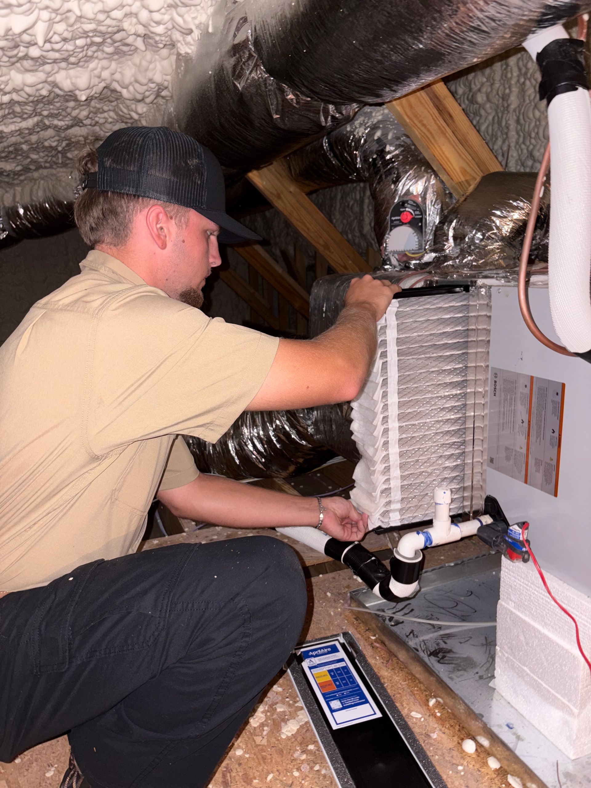 HVAC technician inspecting an air handler in an attic. He wears a hat and khaki shirt.