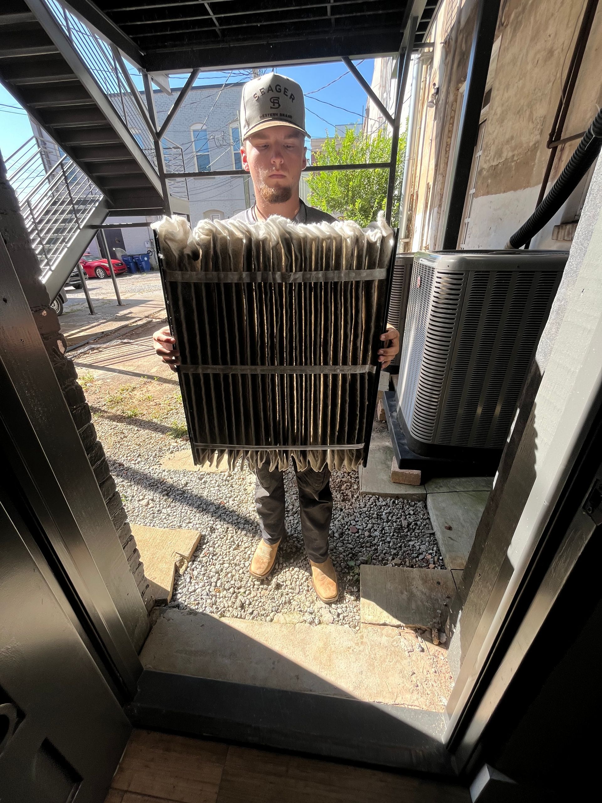 Man holding a large, dirty air filter outdoors, under a metal fire escape.
