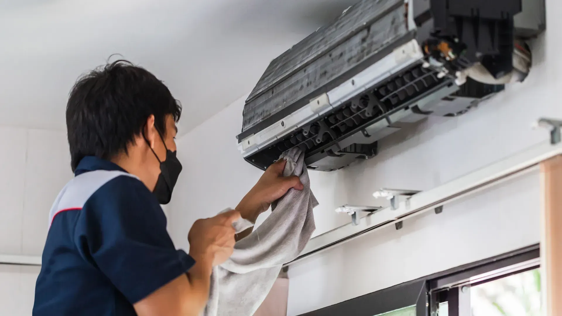A man wearing a mask is cleaning an air conditioner with a towel.