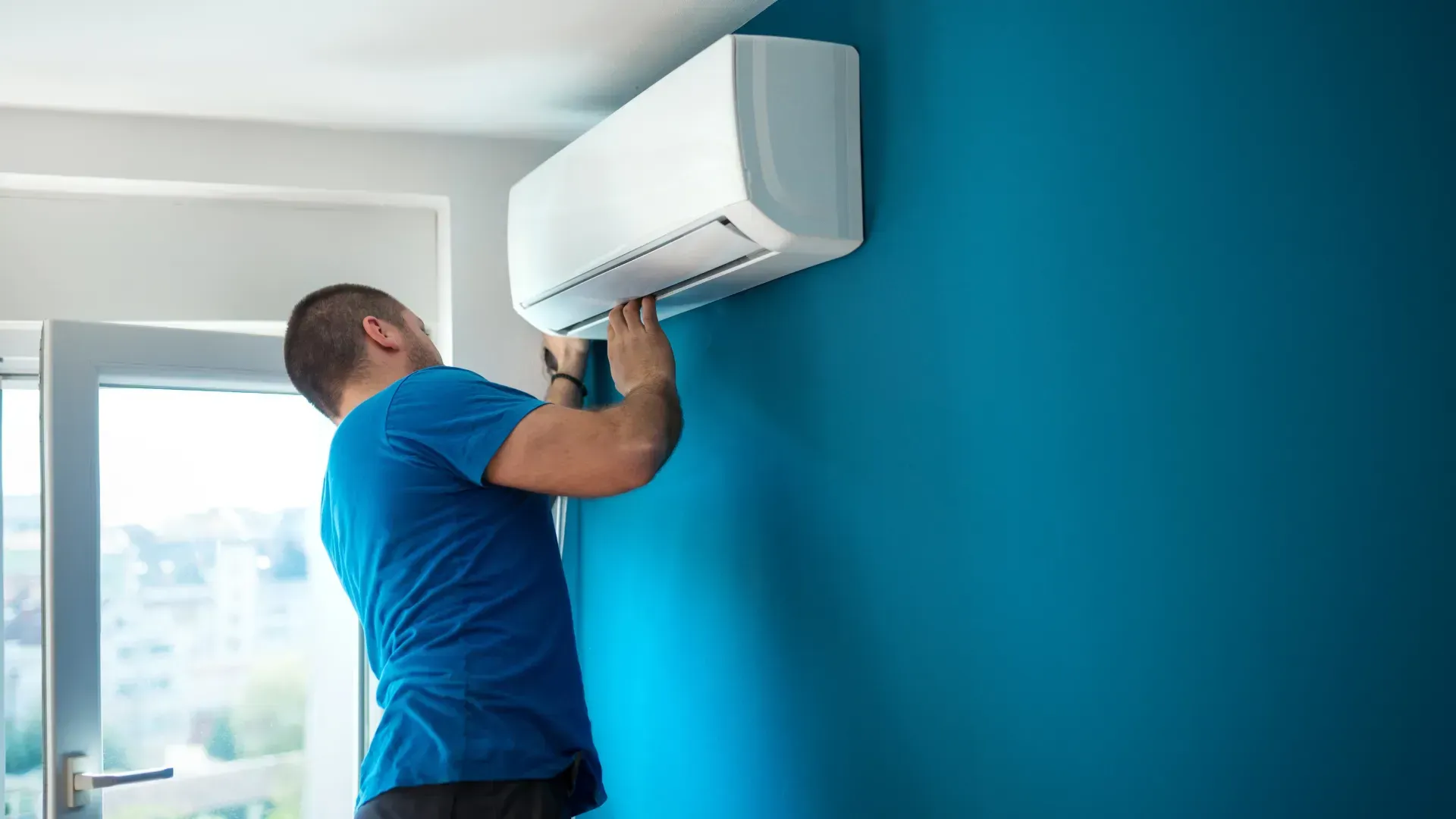 A man is installing an air conditioner on a blue wall.