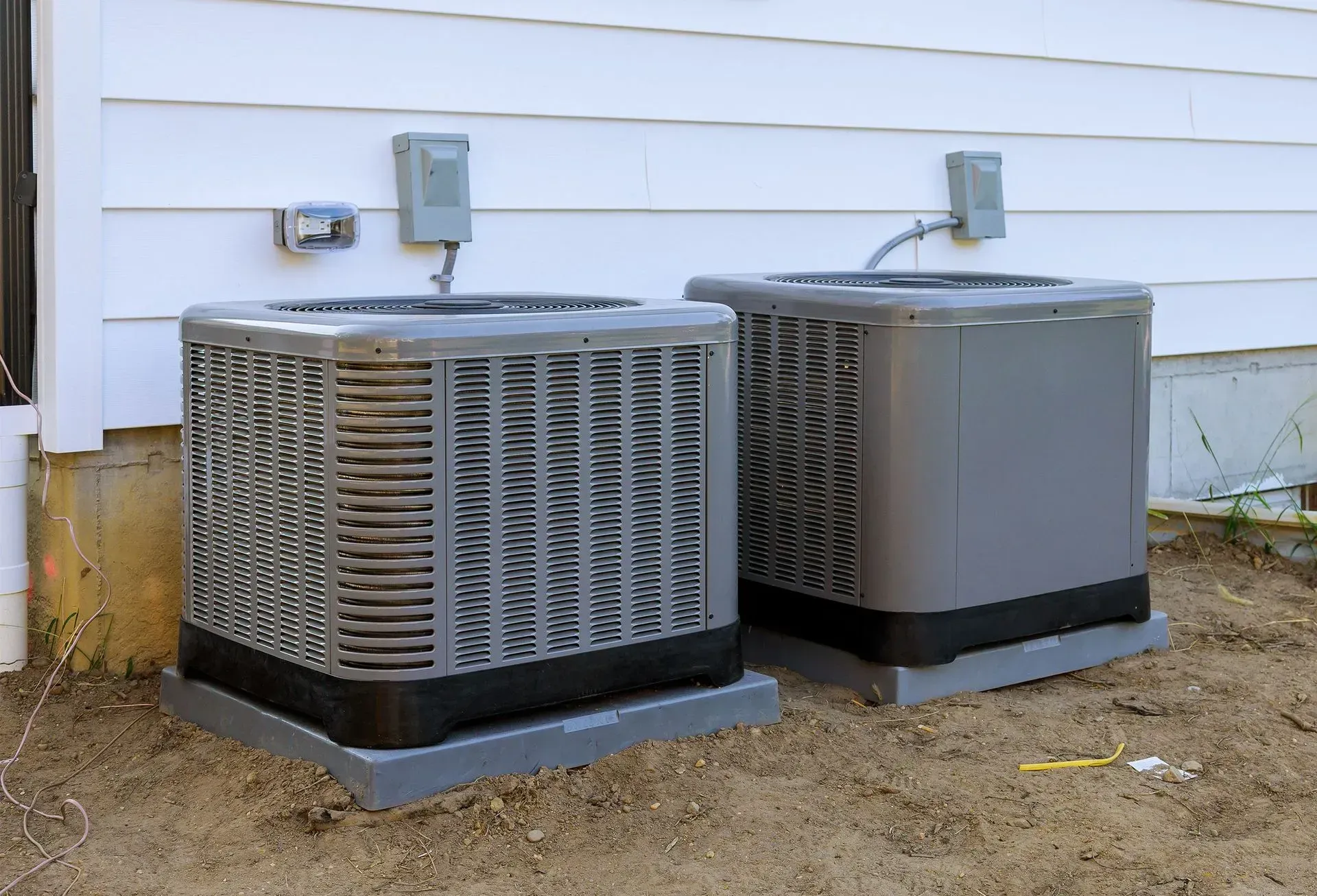 Two air conditioners are sitting next to each other on the side of a house.
