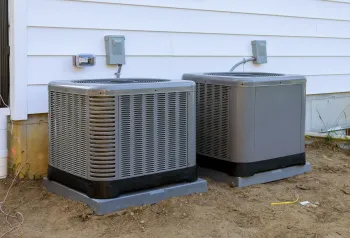 Two air conditioners are sitting next to each other on the side of a house.