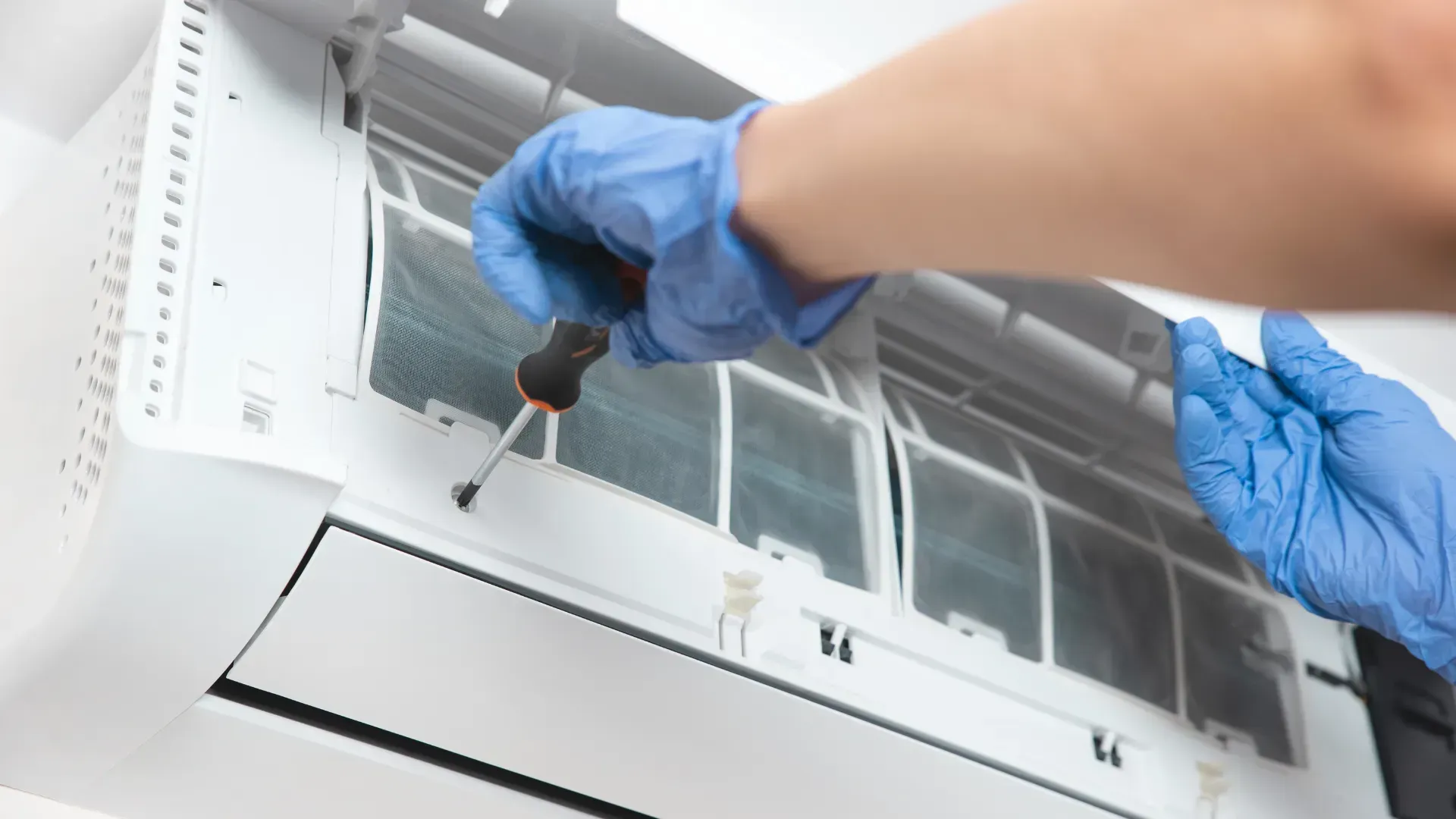 A person wearing blue gloves is fixing an air conditioner with a screwdriver.