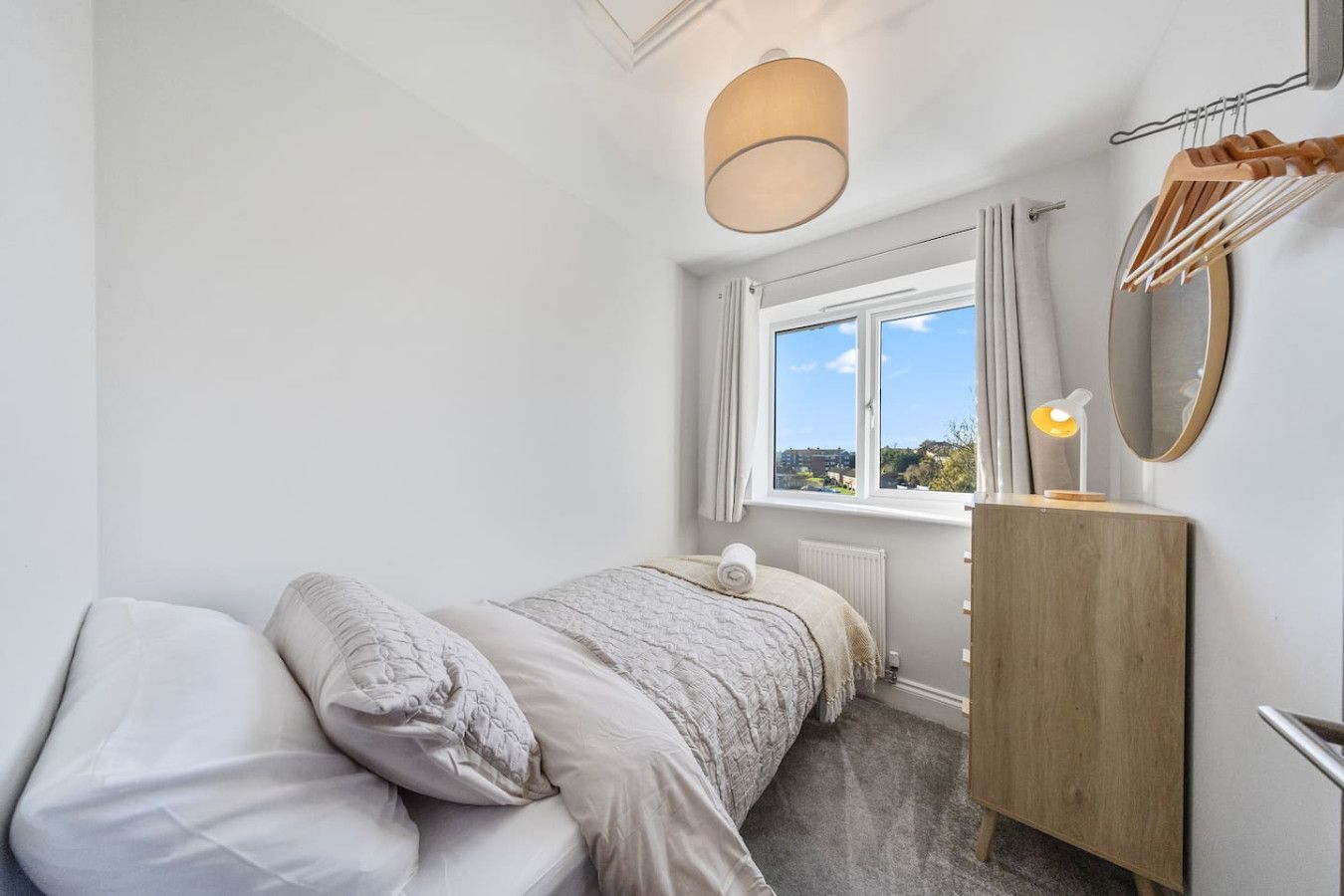 A small, neutral-toned bedroom with a single bed, a window with a landscape view, a wooden chest of drawers, and a mirror.
