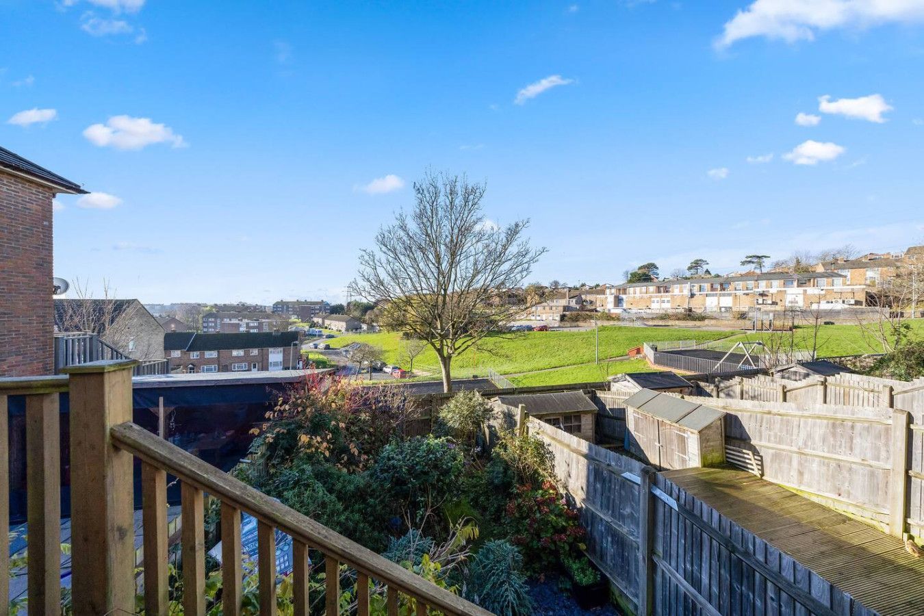 A view from a wooden balcony overlooking a residential garden, a green field, and distant houses under a bright blue sky.