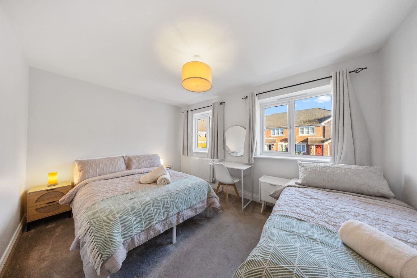 A bedroom featuring a double bed, a twin bed, a wooden nightstand, and a vanity desk under a window with gray curtains.