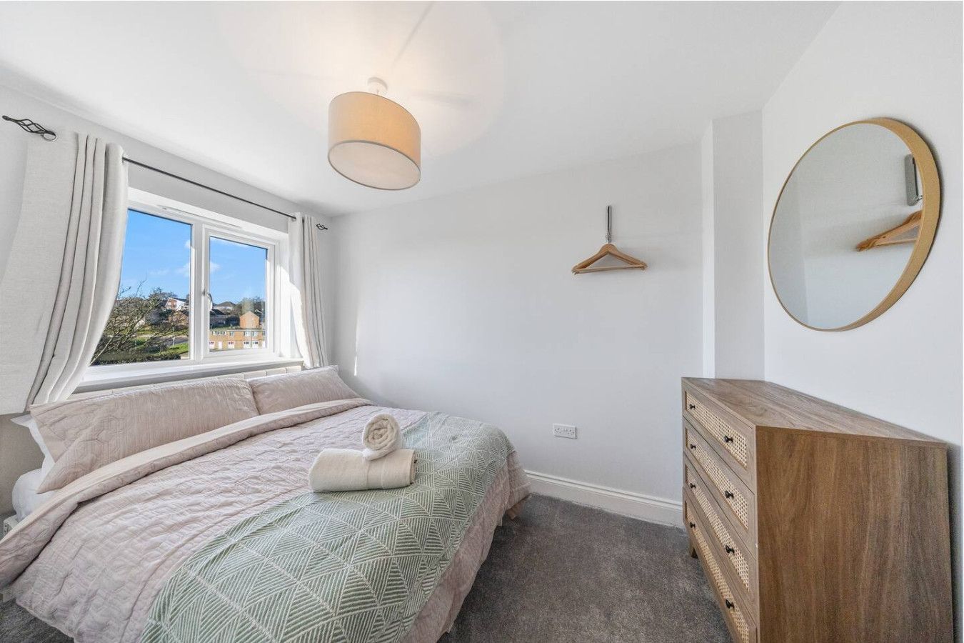 A bright bedroom featuring a queen bed with a patterned cover, a wooden dresser with a round mirror, and a window.